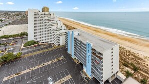 Ein mehrstöckiges Hotel mit Parkplatz vor der Tür, neben einem Strand und dem Meer.