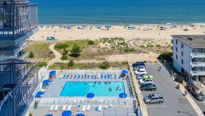 Ein Schwimmbad mit Liegestühlen und Sonnenschirmen, ein Strand mit Sonnenschirmen sowie ein Parkplatz mit Autos.