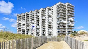 Un edificio residencial de varios pisos con balcones, un sendero de arena que lleva a la entrada y un cartel que dice 'Dune Crossing'.