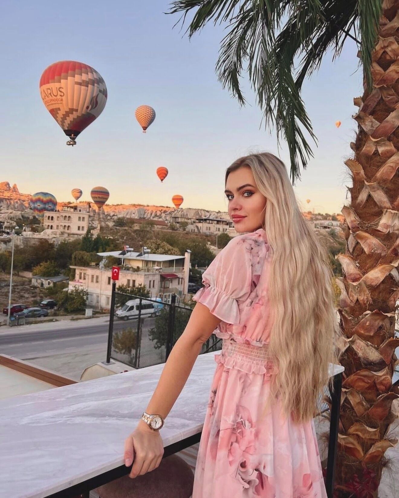 A person standing by a balcony overlooking a cityscape with hot air balloons in the distance.