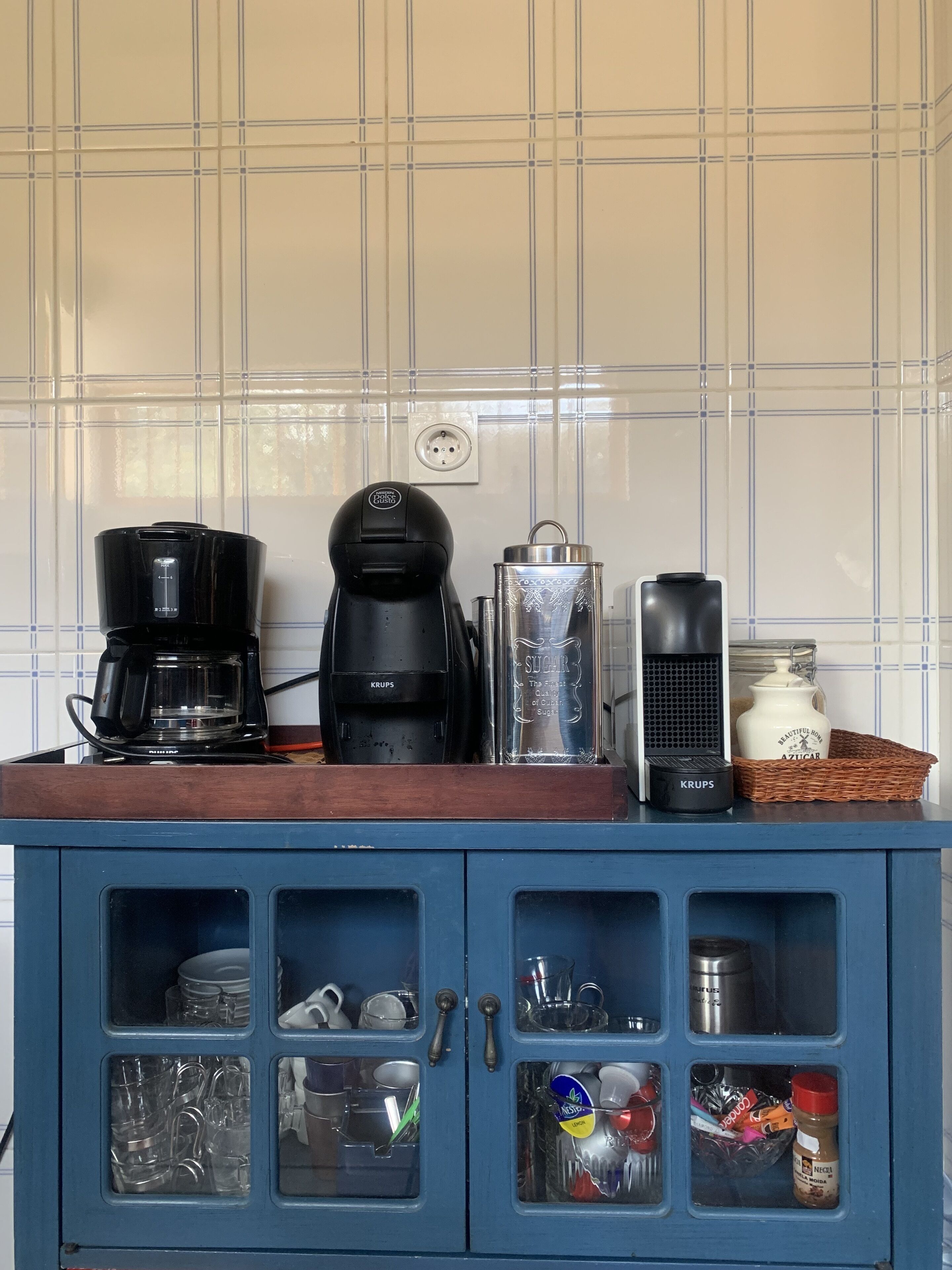 Una encimera de cocina con dos cafeteras, un tarro de azúcar y una cesta. Abajo, se ven armarios con distintos utensilios de cocina.