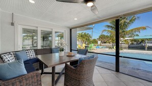 Eine überdachte Terrasse mit Esstisch und Korbmöbeln, große Fenster mit Blick auf einen Swimmingpool und Palmen.