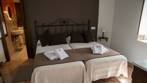 Uma cama de casal com duas almofadas e duas toalhas, uma mesinha de cabeceira com luminária, e um banheiro com pia e espelho.