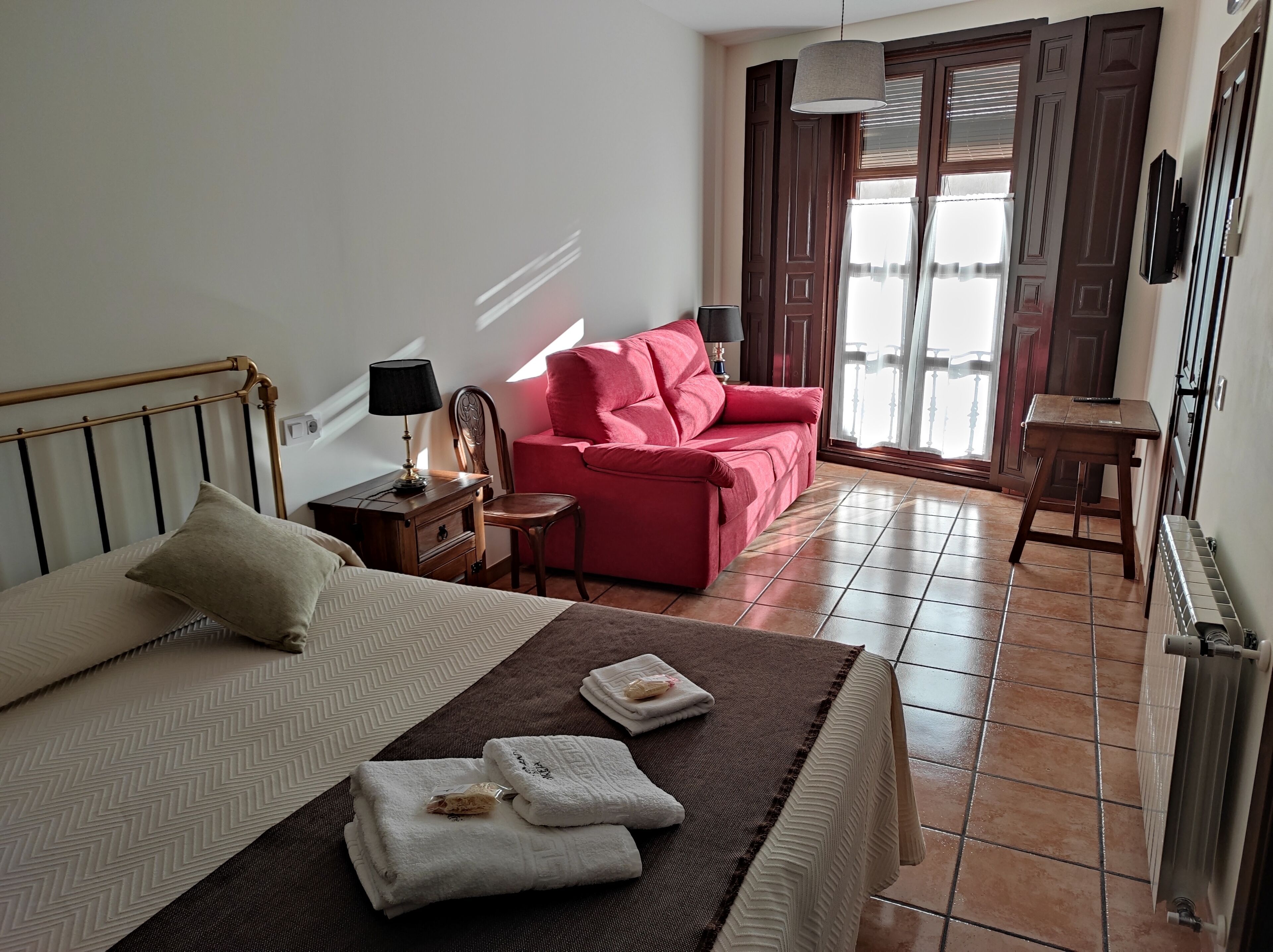 A hotel room with a bed, a red armchair, a small table with a lamp, and a view of the outside through a window.