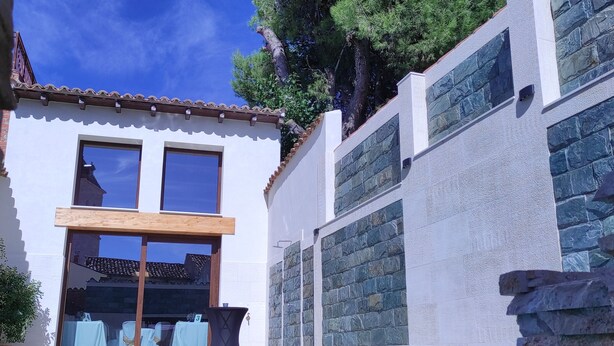 Uma área à beira da piscina com céu azul límpido, um edifício branco com grandes janelas e um muro de pedra.