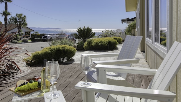 En træterrasse med hvide stole, et bord med et vinglas og druer, samt havudsigt.