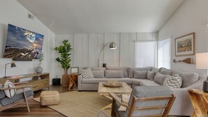 En moderne stue med en hjørnesofa, et sofabord, en træstol, et fjernsyn og en stor plante.