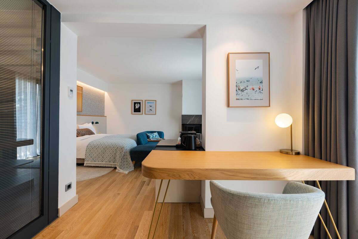 A modern hotel room with a wooden desk, a comfortable chair, a bed with a blue headboard, and a framed picture on the wall.
