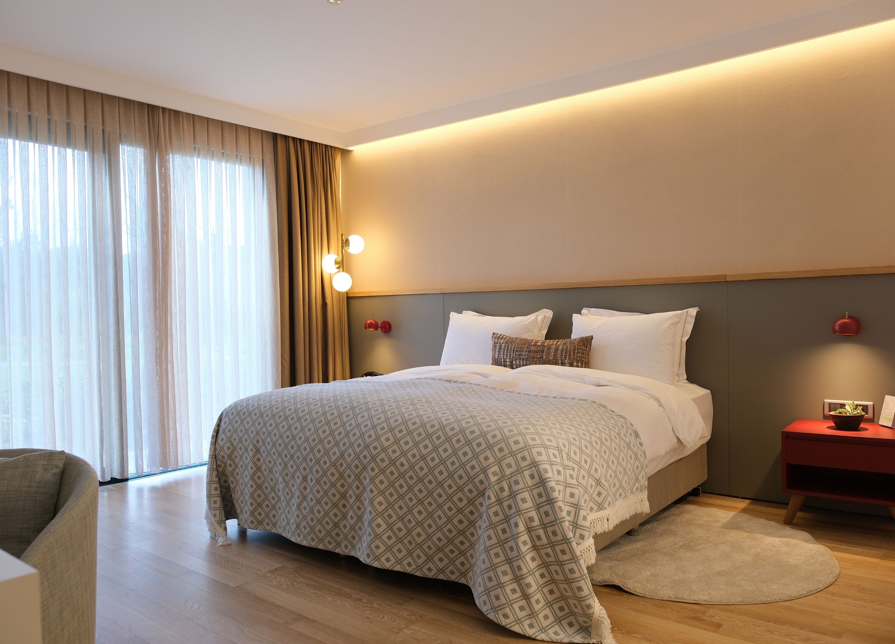 A modern hotel room with a large bed, a bedside table with a red lamp, and a view of the outdoors through sheer curtains.