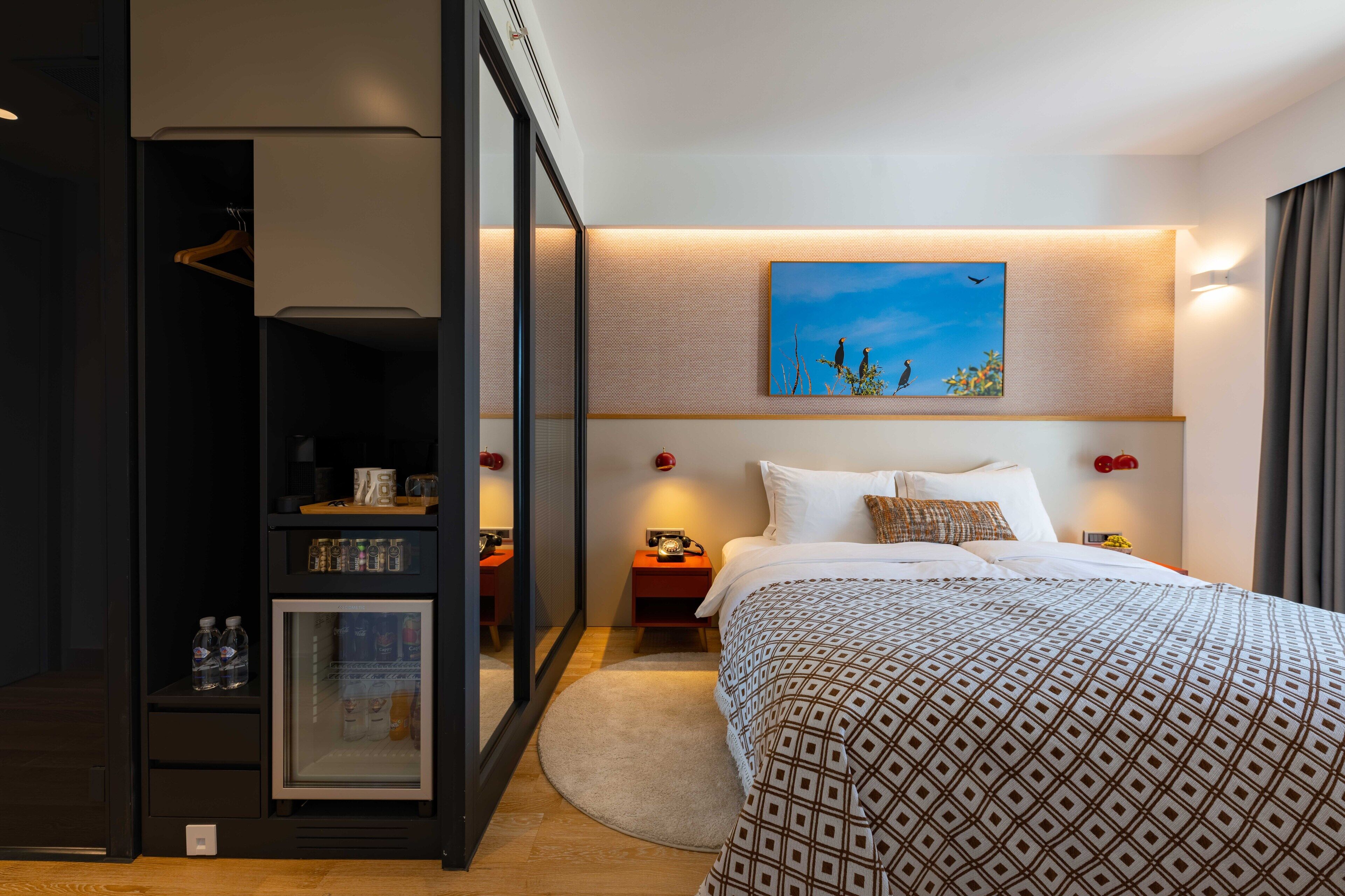 A modern hotel room with a large bed, a bedside table with a lamp, a nightstand with a telephone, and a wardrobe with a mirror.