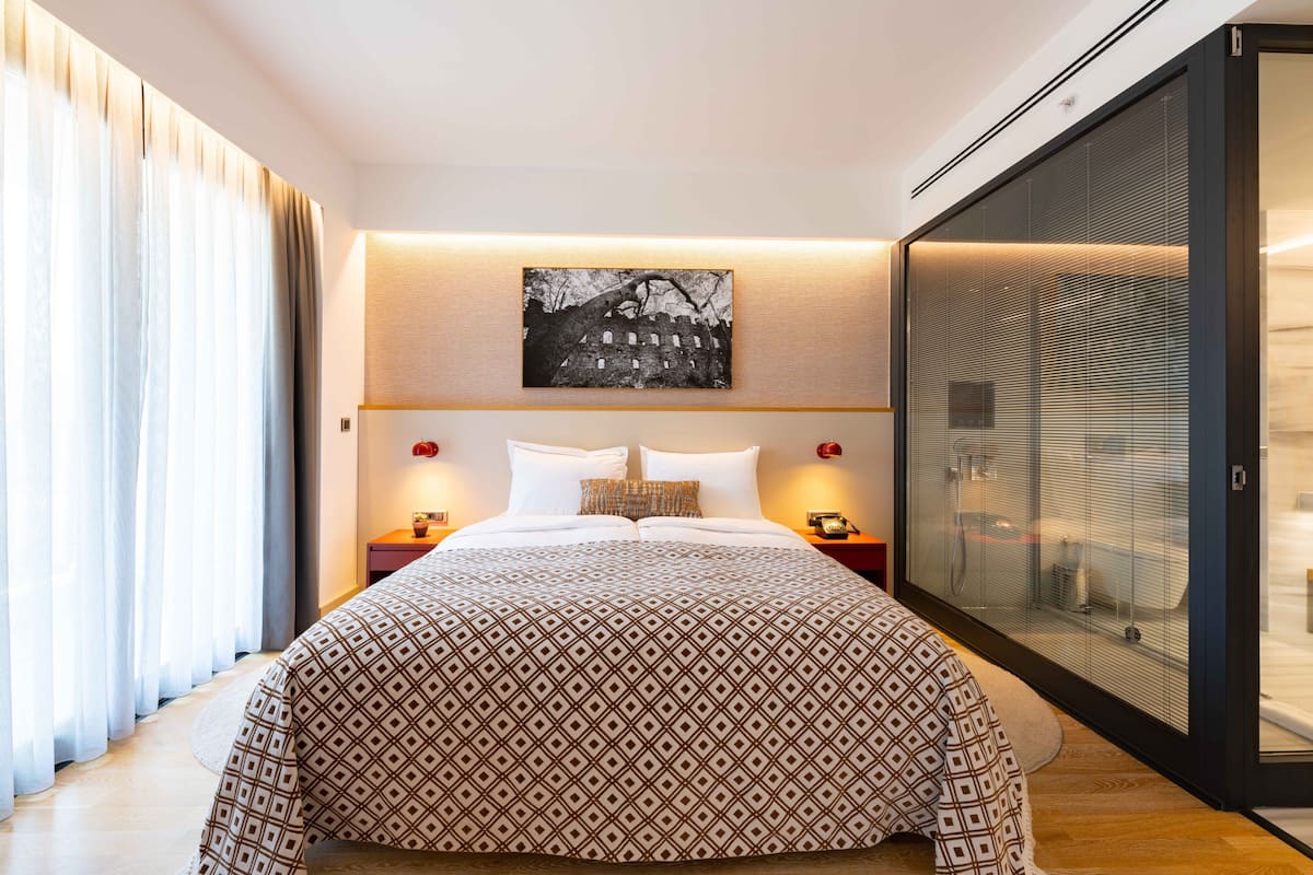 A modern hotel room with a large bed, a glass partition, and a cityscape artwork on the wall.