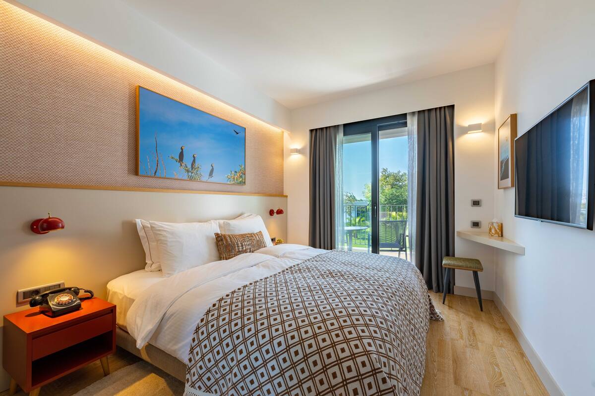 A modern hotel room with a large bed, a nightstand with a telephone, a painting on the wall, a window with curtains, and a view of greenery outside.