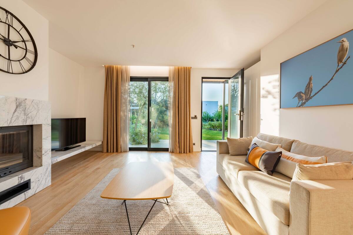 A modern living room with a fireplace, a flat-screen TV, a sofa, and a large window with curtains.