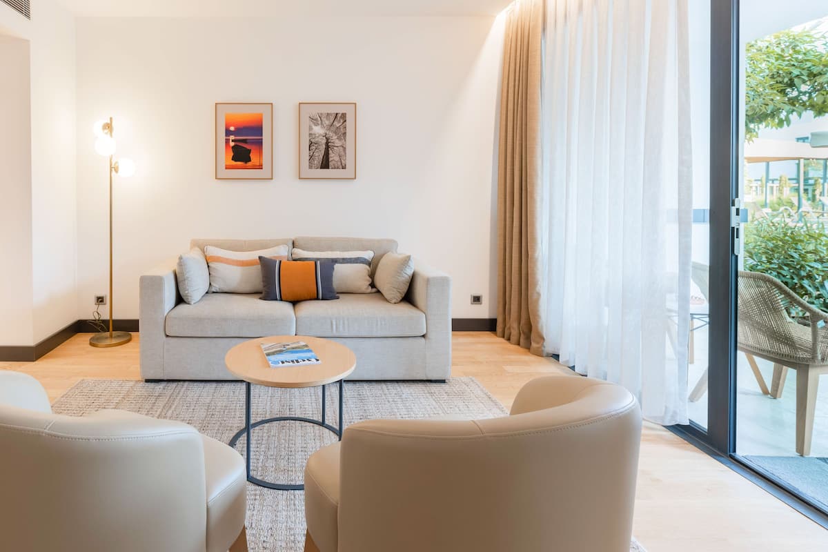 A modern living room with a sofa, two armchairs, a coffee table, and a floor lamp. There are two framed pictures on the wall and a large window with curtains.