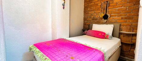 A room with a brick wall, a bed with a white mattress and pink bedding, and a small wooden side table.