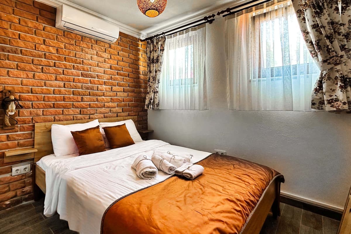 A hotel room with a brick wall, a bed with white bedding and brown pillows, a window with floral curtains, and a wall-mounted air conditioner.