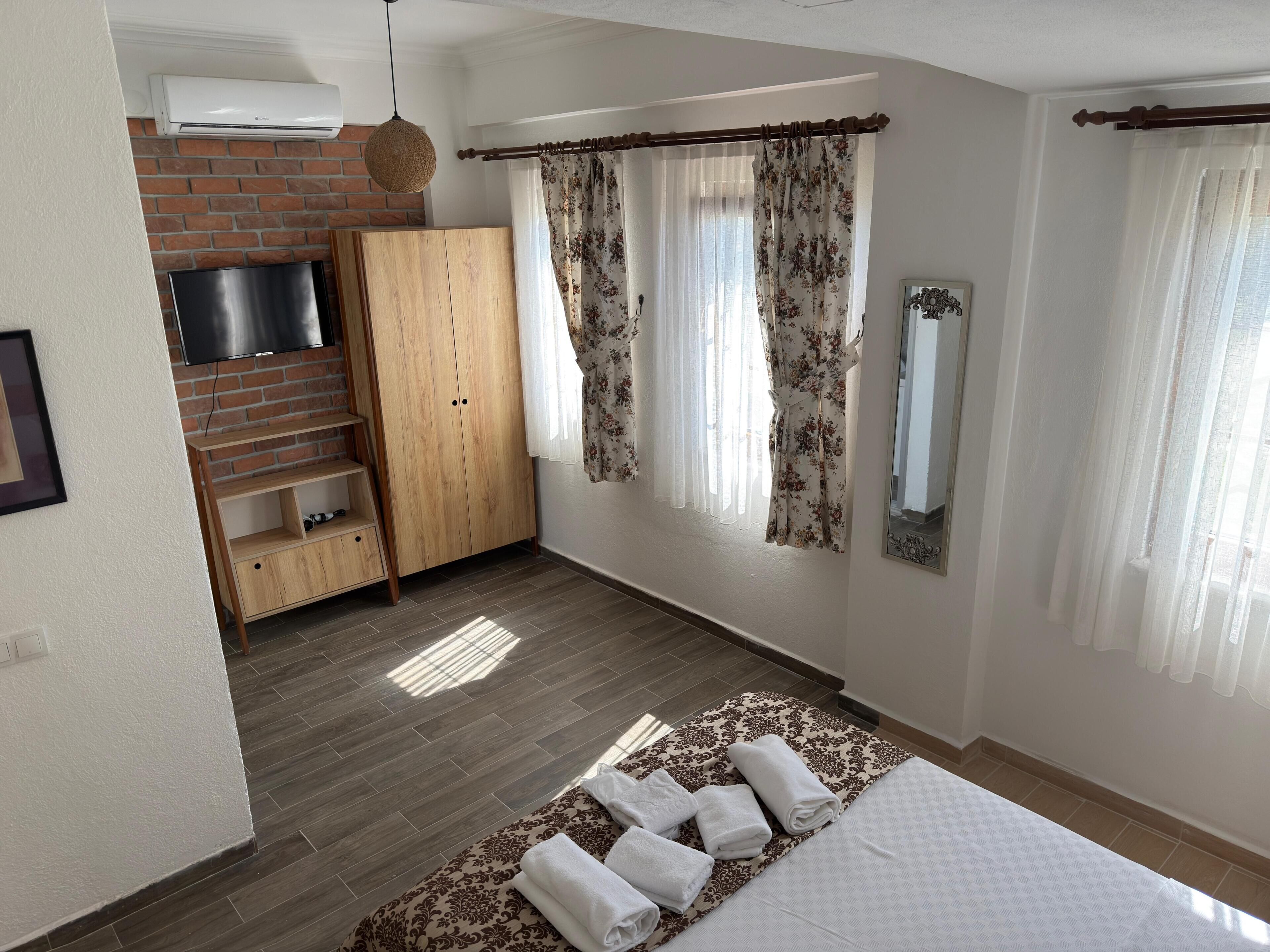 A bedroom with a brick wall, a TV, a wardrobe, a bed with towels, and a window with curtains.