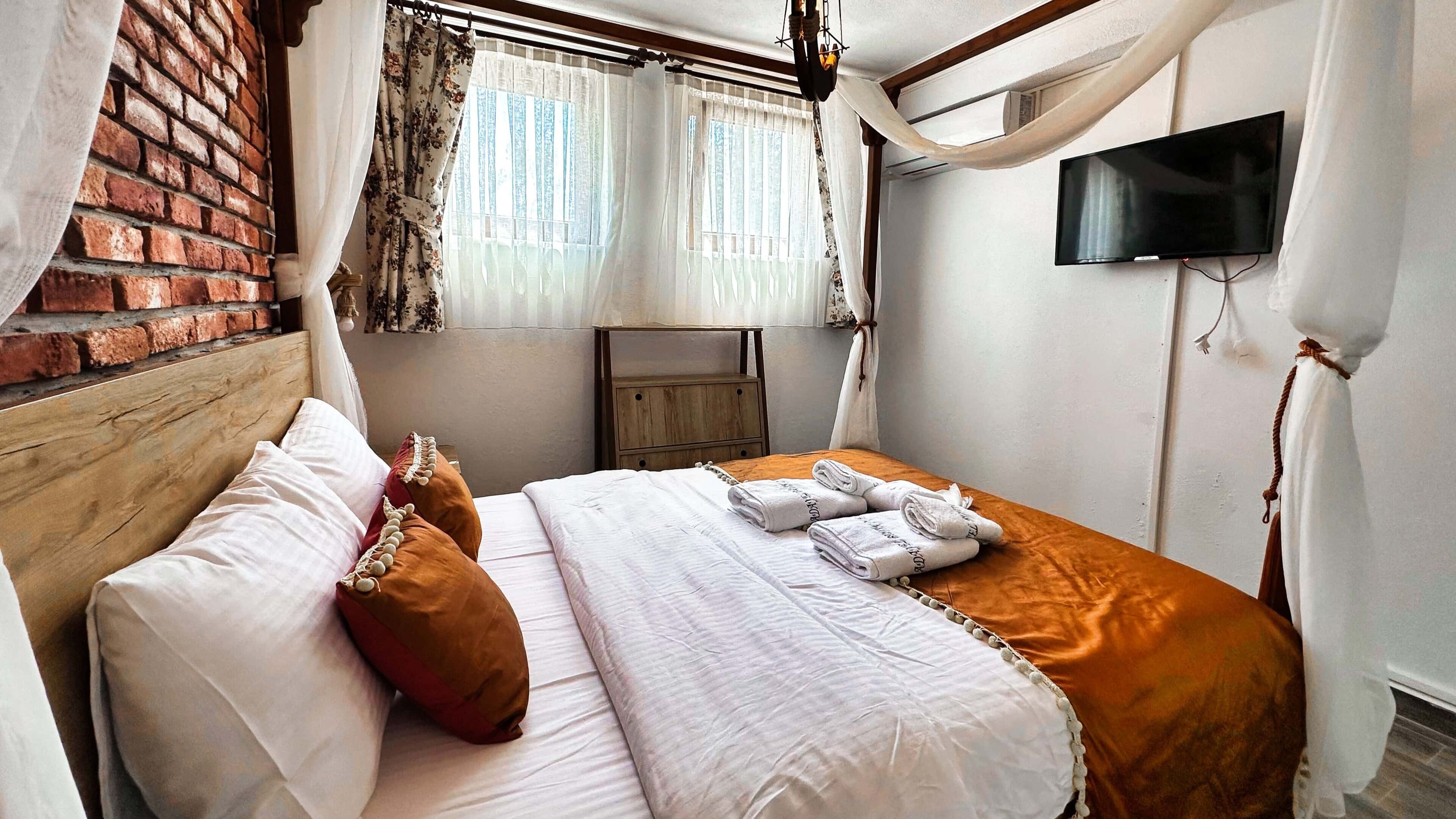 Un dormitorio con pared de ladrillo, cabecera de madera, cama con sábanas blancas y almohadas marrones, mesita de noche de madera, televisor montado en la pared y ventana con cortinas de gasa.
