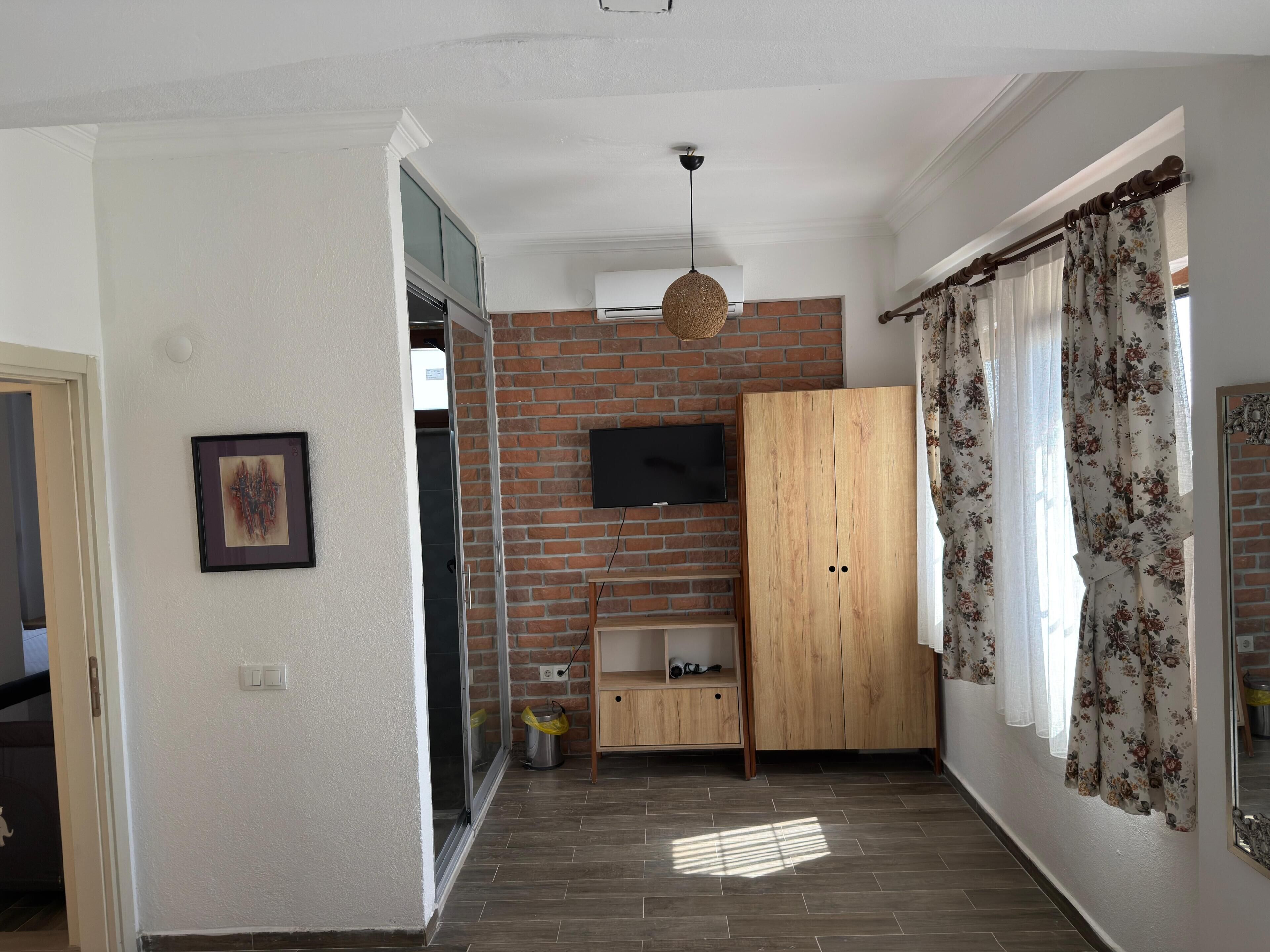 A room with a brick wall, a wooden wardrobe, a television, and a window with floral curtains.