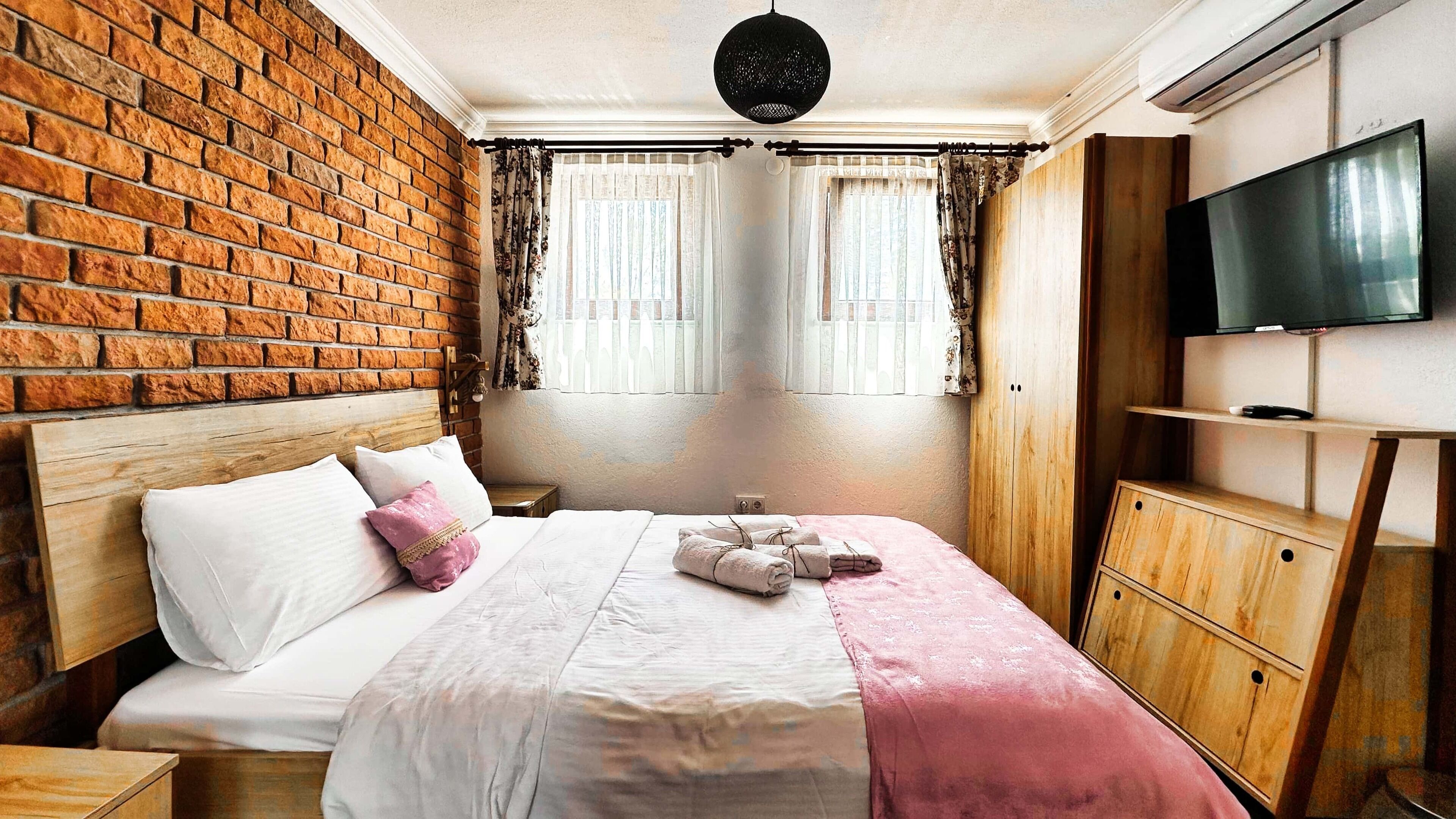 A hotel room with a brick wall, a bed with white and pink bedding, a wooden headboard, a wooden wardrobe, and a television on the wall.