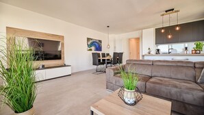 Una sala de estar moderna con un televisor de pantalla plana grande, un sofá de cuero marrón y una mesa de centro de madera. Se observa un área de comedor con una mesa y sillas al fondo.
