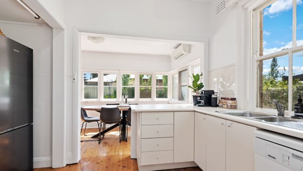 A modern kitchen with white cabinetry, a built-in dishwasher, and a dining area with a table and chairs. - Heritage on Hill - Refreshing Elegance (Orange)