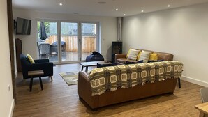 A living room with a brown sofa, a blue armchair, and a coffee table. There is a sliding glass door leading to a patio area with outdoor furniture. - Pennant Uchaf (Llanbrynmair)
