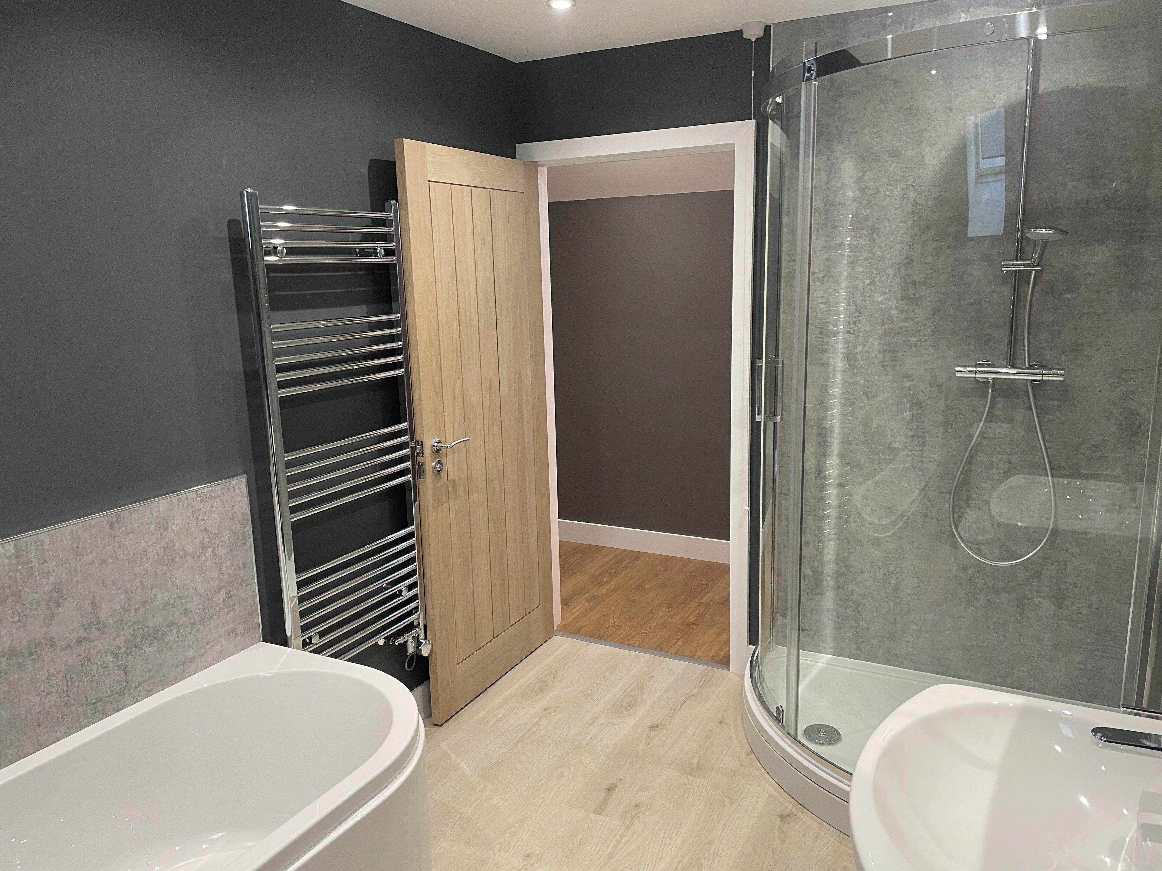 A modern bathroom with a glass-enclosed shower, a white bathtub, and a wooden door leading to a walk-in closet.
