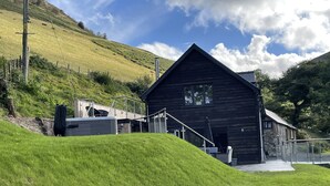 A rustic wooden building with a steep roof, a staircase leading to the entrance, and a grassy hill in the background. - Pennant Uchaf (Llanbrynmair)