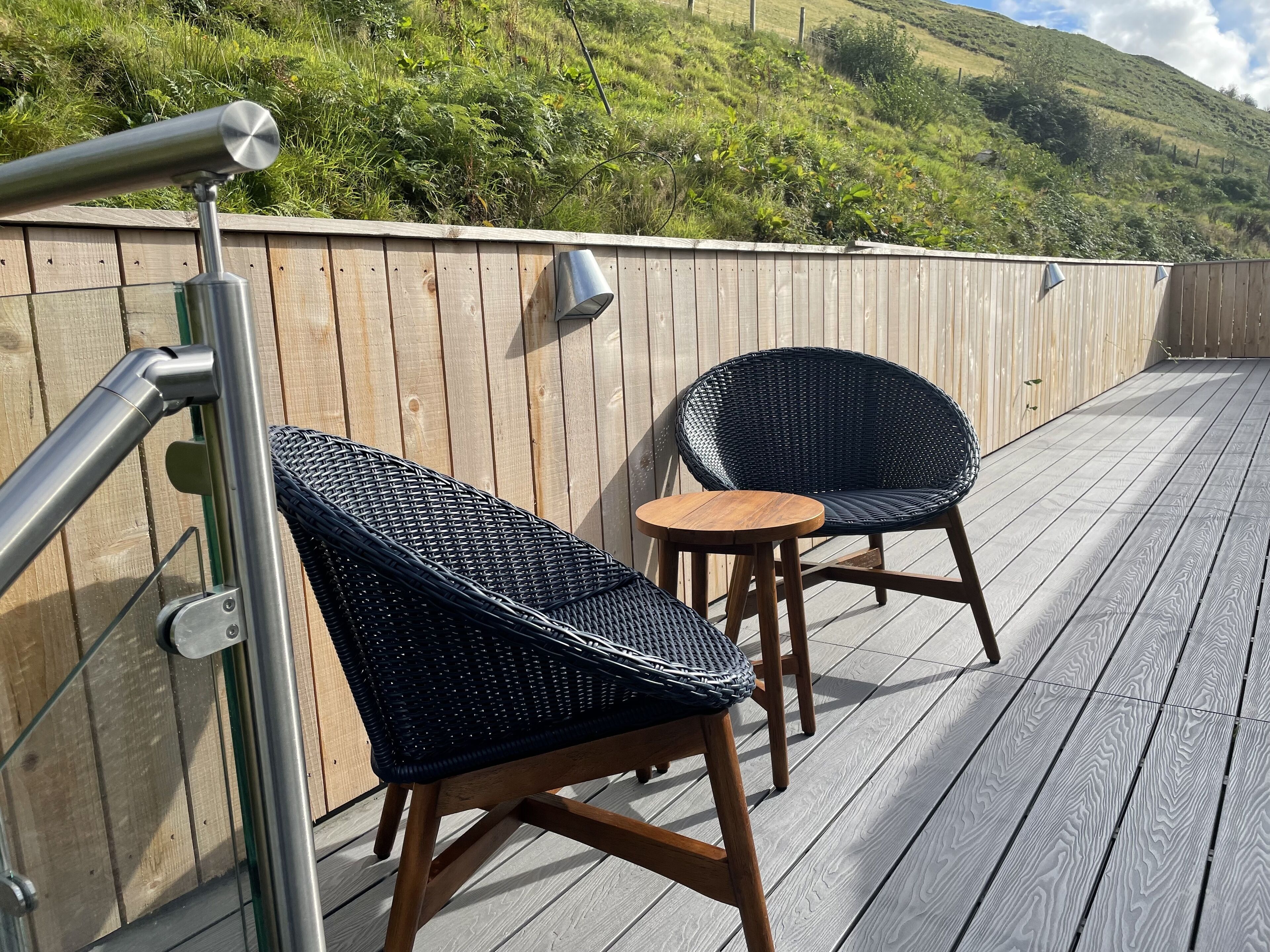 A deck with two wicker chairs and a small table, overlooking a lush green hillside.
