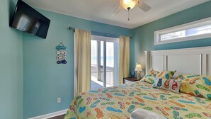 A bedroom with a bed, a ceiling fan, a TV mounted on the wall, and a window with curtains. - Shore Thing | Oceanfront + Private Beach Access! (Surf City)
