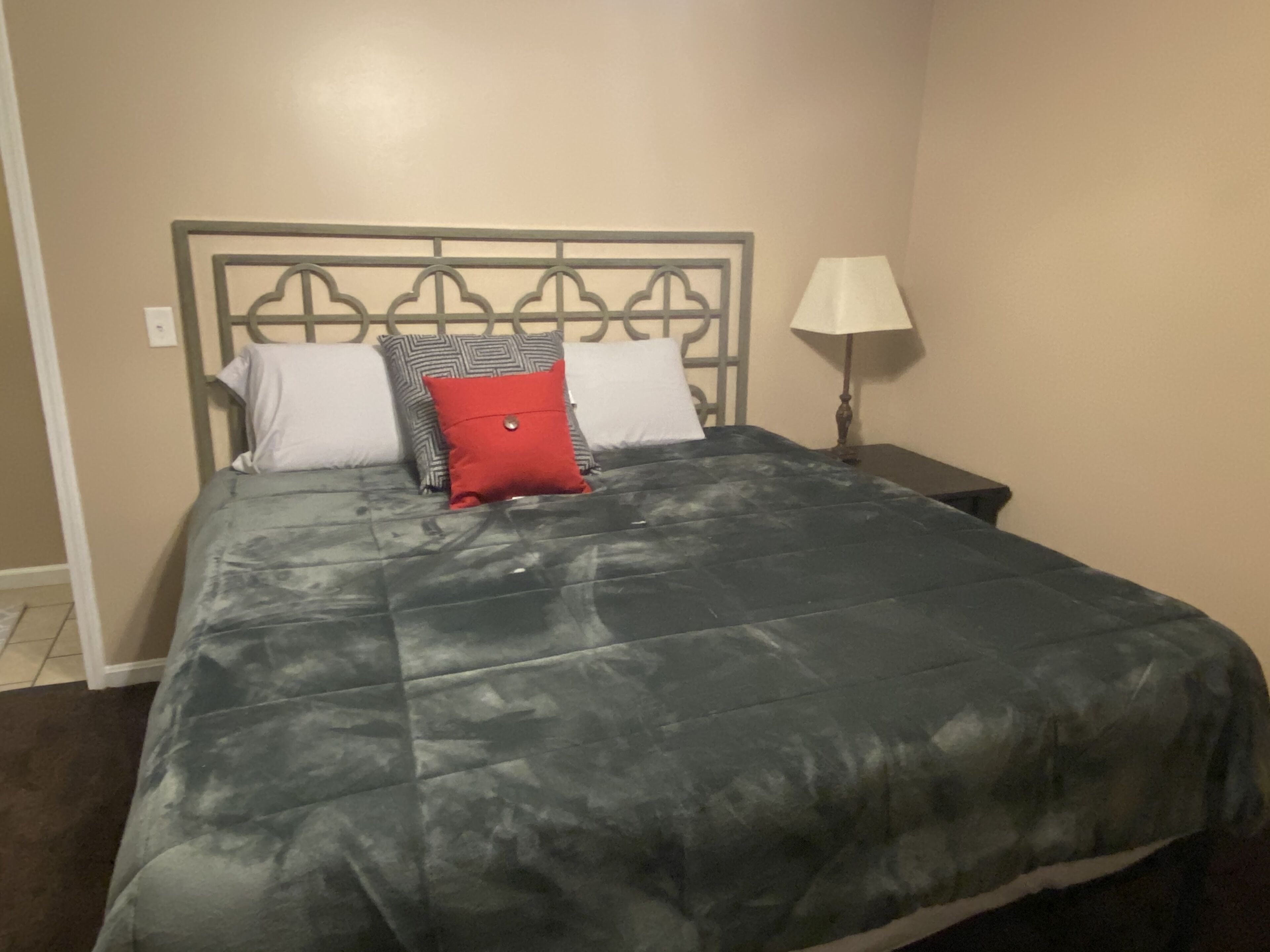 A bed with a grey quilt, two white pillows, and a red pillow. There is a nightstand with a lamp to the right.