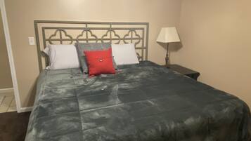 A bed with a grey quilt, two white pillows, and a red pillow. There is a nightstand with a lamp to the right.