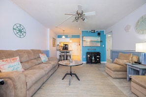 A living room with a sofa, a coffee table, and a dining area with chairs. There is a ceiling fan, a fireplace, and a painting on the wall.