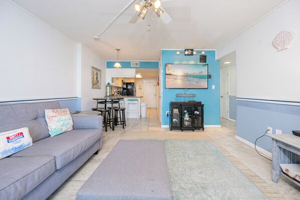 A living room with a gray sofa, a coffee table, and a dining area with a kitchen in the background. - Gulf Shores Surf & Racquet 202B | Lagoon Views (Gulf Shores)