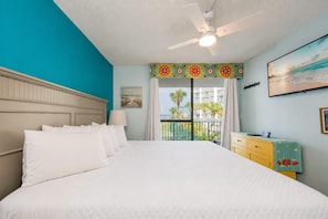 A hotel room with a large bed, a ceiling fan, a view of the ocean, and a balcony with a railing.