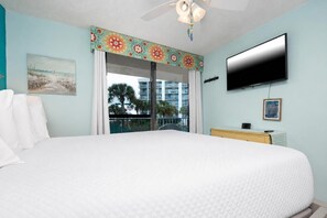 A hotel room with a bed, a television, a balcony with a view of palm trees and buildings, and a ceiling fan. - Gulf Shores Surf & Racquet 202B | Lagoon Views (Gulf Shores)