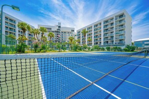 A tennis court with a net in the foreground, surrounded by a fence, and a multi-story hotel building in the background.