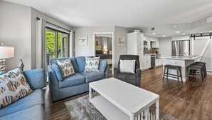 A modern living room with a sofa, armchair, and coffee table, a kitchen with white cabinets and stainless steel appliances, and a large window offering a view of greenery. - Coastal 2BR Villa w/ Balcony & Amenities (Hilton Head Island)