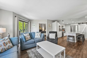 Uma sala de estar moderna com sofá, poltrona e mesa de centro, uma cozinha com armários brancos e eletrodomésticos de aço inoxidável, e uma janela grande com vista para o verde.
