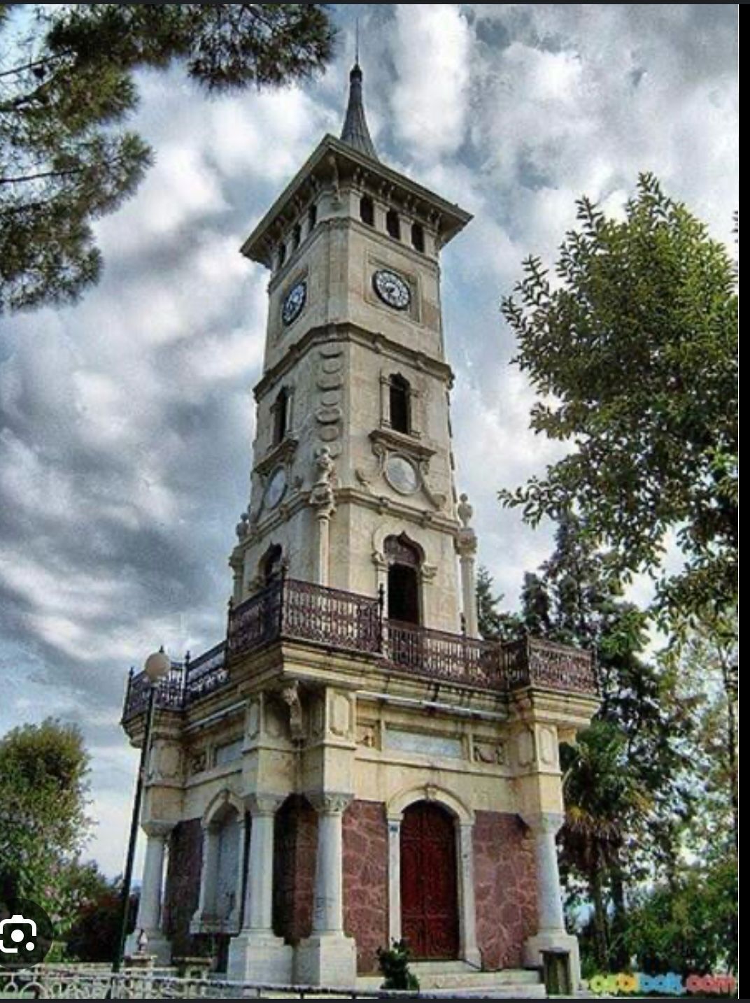 Uma torre de relógio histórica com varanda, rodeada por árvores.