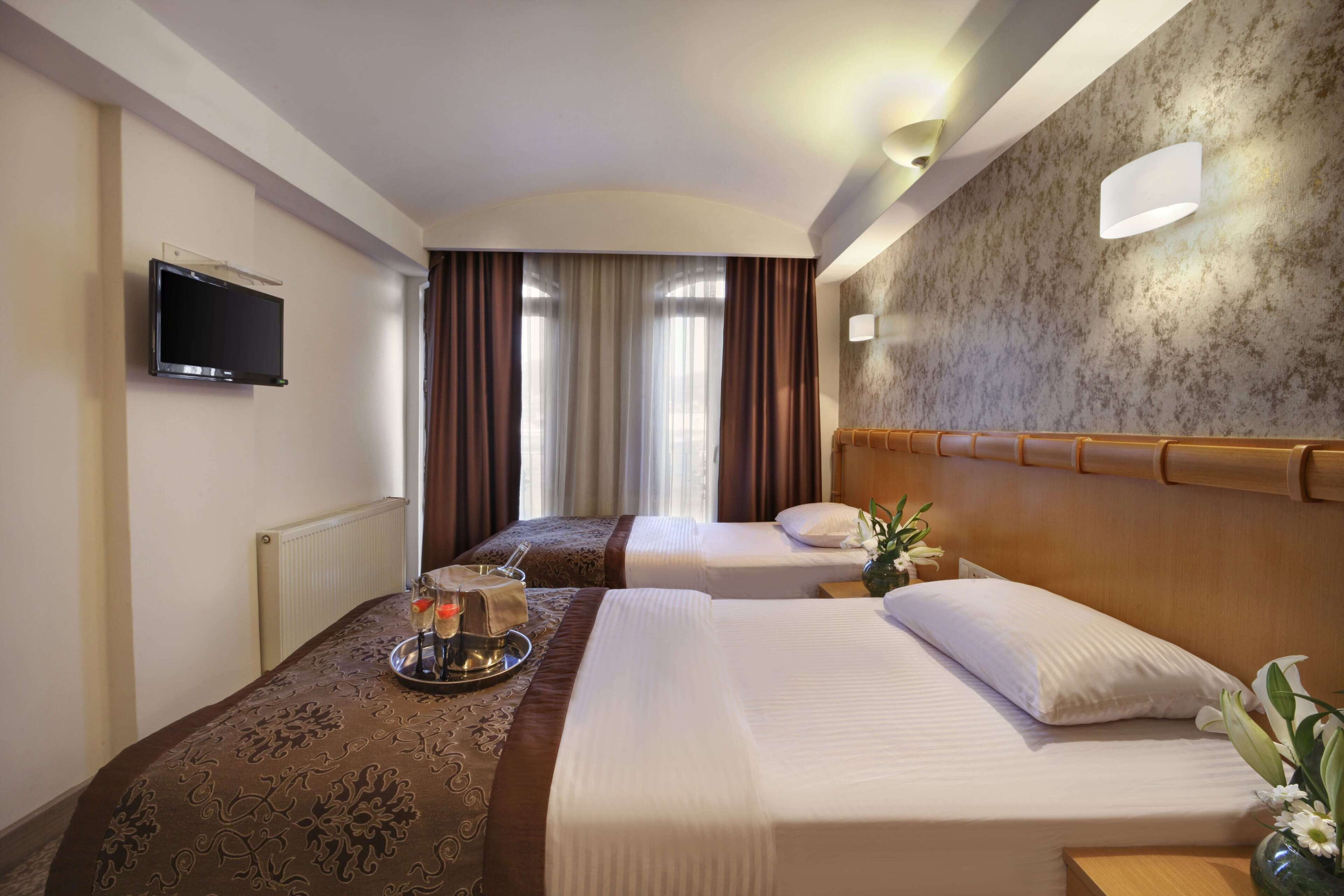 A hotel room with two beds, a flat-screen TV, a tray with a candle and a glass, and a vase with flowers on the nightstand.