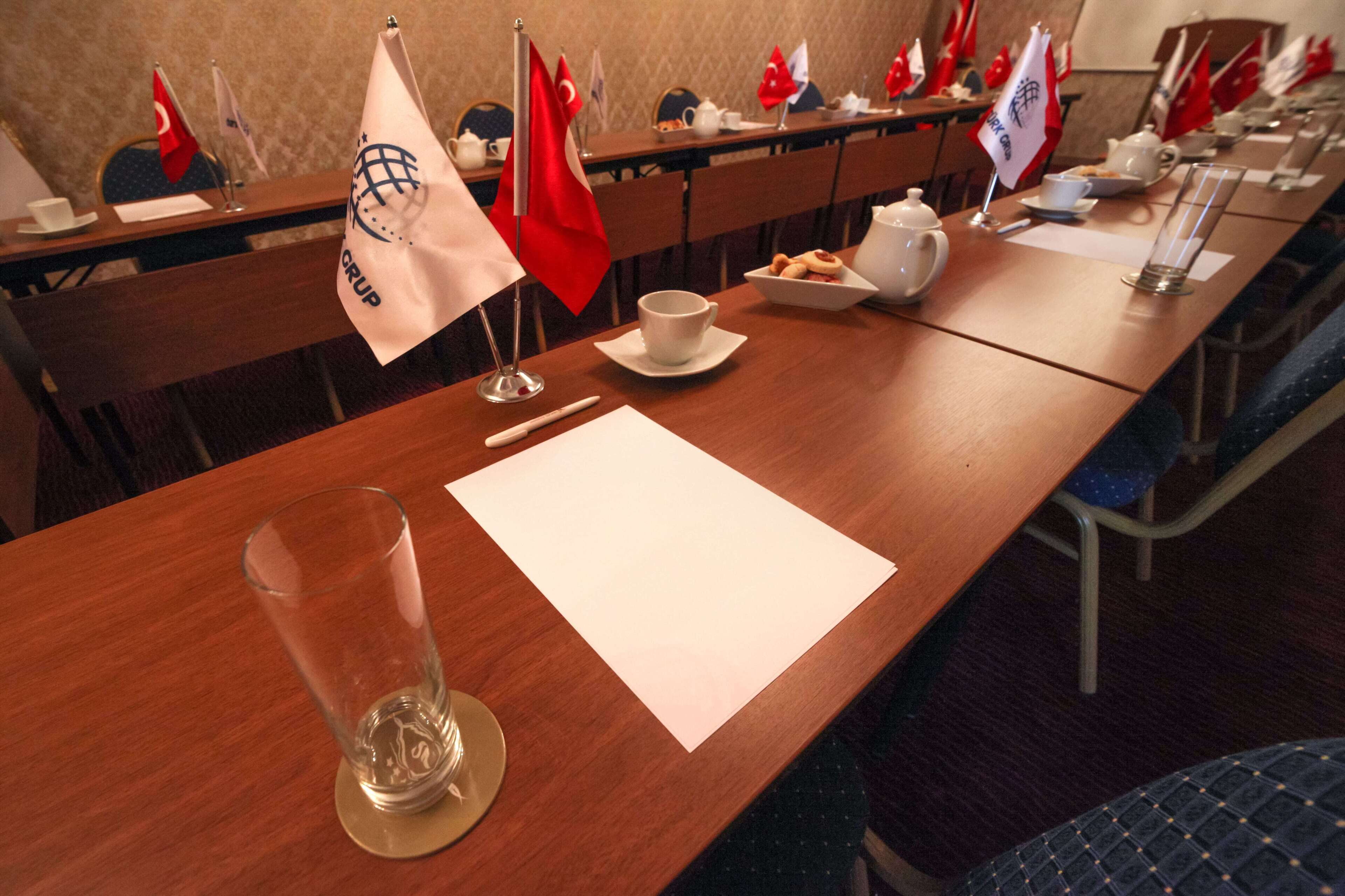 A conference room with a long table set for a meeting, flags of various countries, and a glass of water on the table.