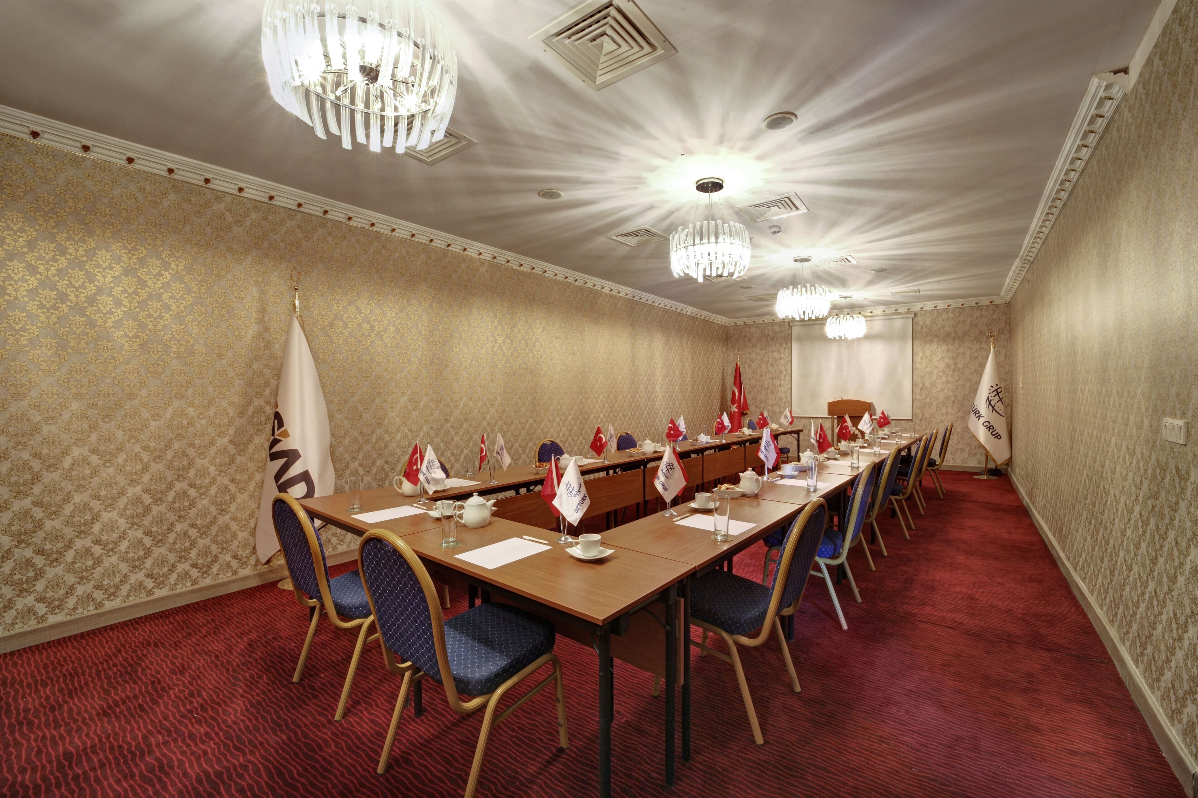 Uma sala de conferência com uma mesa comprida, cadeiras, bandeiras e lustres.