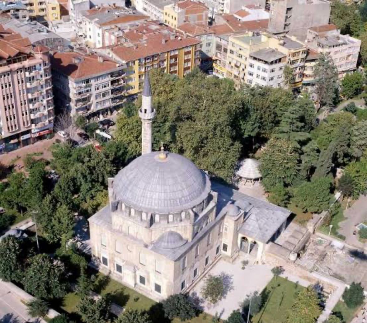 Vista aérea de un edificio histórico con una cúpula destacada y un minarete, rodeado de abundante vegetación y edificios urbanos colindantes.