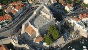 Vista aérea de una fortificación histórica con edificios circundantes, una carretera pavimentada y un puerto deportivo con barcos.