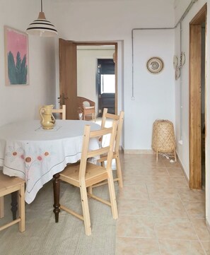 Uma sala de jantar com uma mesa redonda, cadeiras de madeira, toalha de mesa branca, luminária pendente e uma cesta de palha.