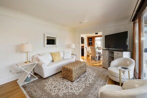 Una sala de estar moderna con un sofá blanco, una alfombra estampada, un sillón blanco, una mesita auxiliar y un televisor de pantalla plana.