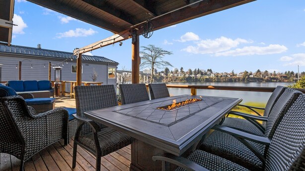 Une terrasse extérieure couverte, dotée d’un foyer central, de meubles en osier et offrant une vue sur un lac et des bâtiments situés au loin.