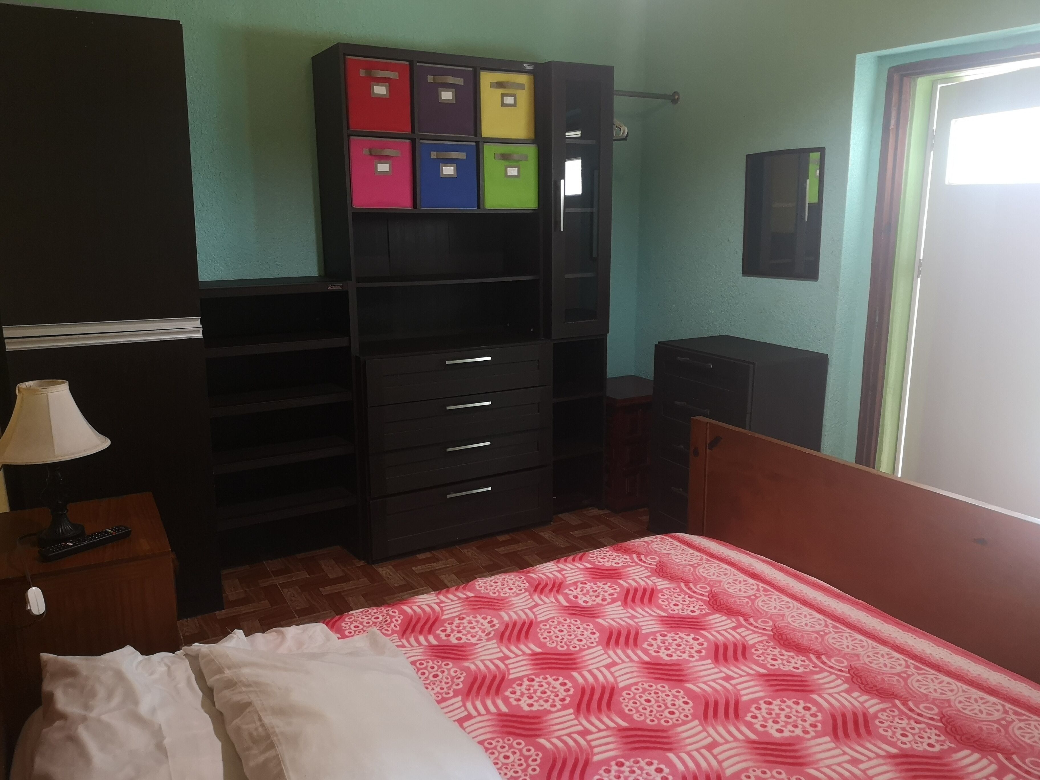 Ein Schlafzimmer mit Bett, Schreibtisch mit Lampe, Kleiderschrank mit Schubladen und einem an der Wand befestigten Fernseher.